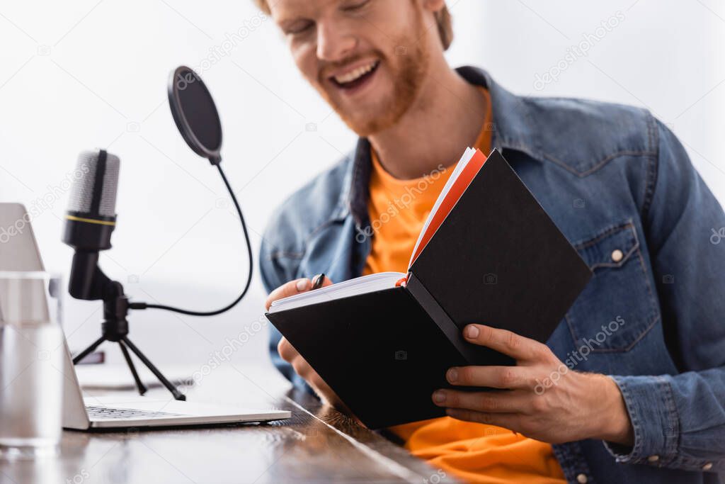 selective focus of young excited broadcaster holding notebook while speaking in microphone