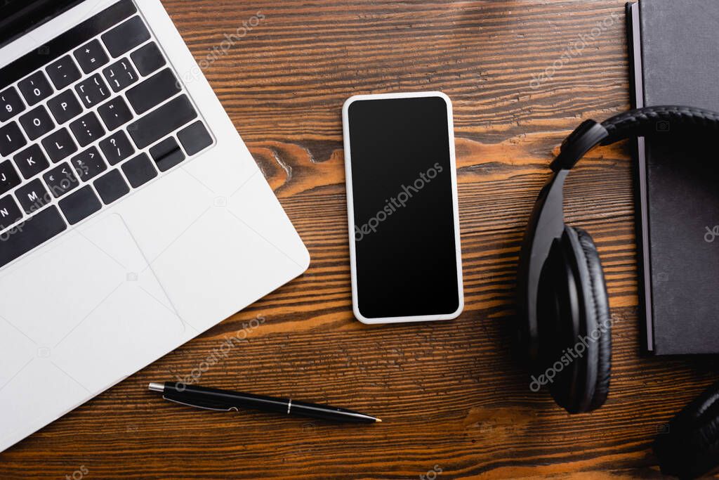 top view of smartphone with blank screen, laptop, wireless headphones, notebook and pen on office desk