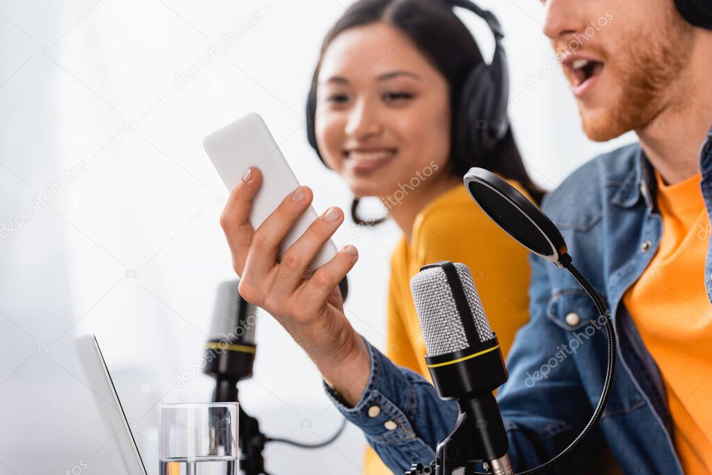 excited announcer using smartphone near asian coworker in wireless headphones