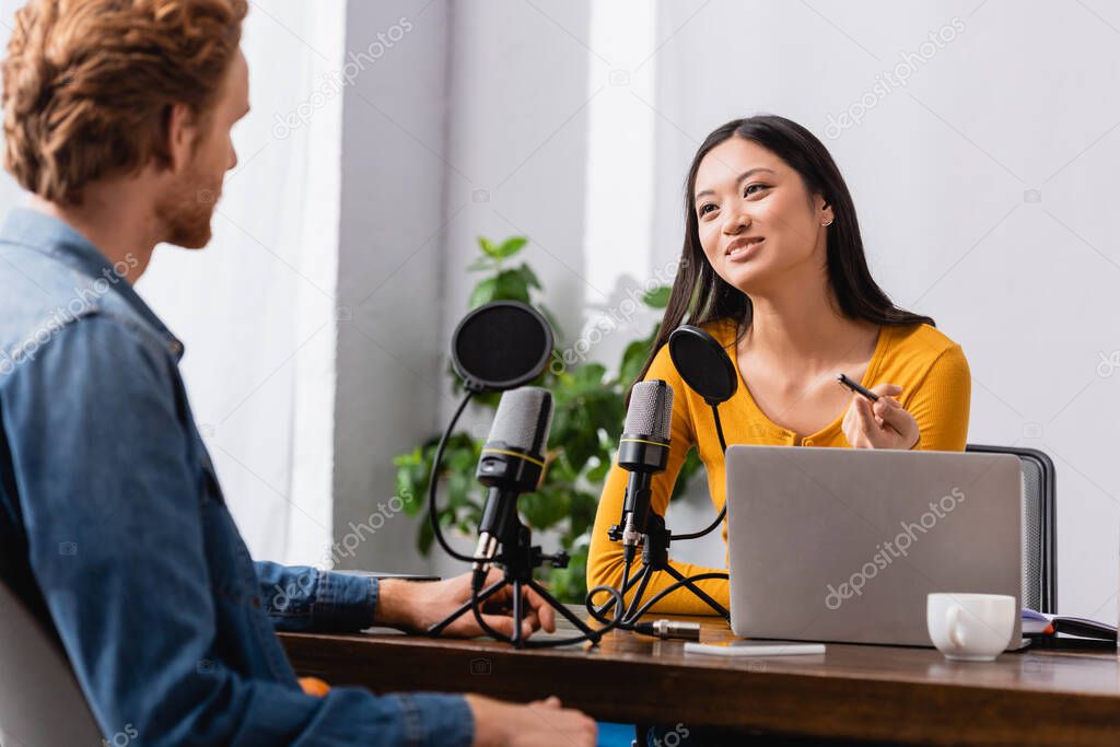 selective focus of young asian interviewer holding pen while talking to redhead man in radio studio