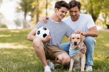 Babası genç oğlunun yanında oturmuş futbol oynuyordu Golden Retriever oynuyordu.