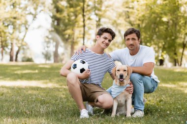 Baba, genç oğlanın yanında oturmuş futbol ve golden retriever oynuyor.