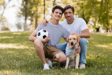 Baba çimlerin üzerinde oturan mutlu genç oğlu futbol ve golden retriever ile