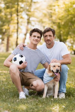 Baba genç çocuğun yanında çimlerde oturuyor futbol ve golden retriever ile