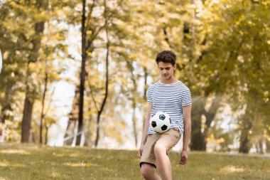 Genç çocuk parkta ağaçların yanında futbol oynuyor. 