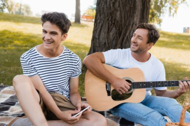 Babasının yanında akıllı telefon tutan genç bir çocuğun akustik gitar çalmasının seçici odağı.