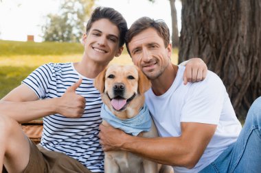 Genç çocuk başparmağını gösteriyor ve Golden Retriever yakınlarında babasına sarılıyor.