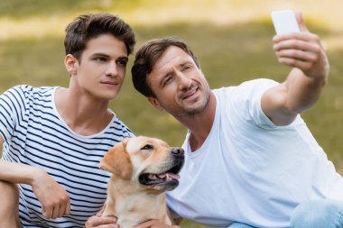 Baba ve genç çocuk parkta Golden Retriever ile selfie çekiyorlar.