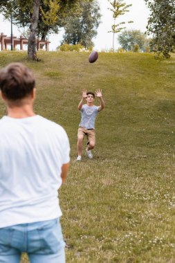 Genç oğlanın parkta babasının yanında rugby topu yakalamasına odaklanmış. 