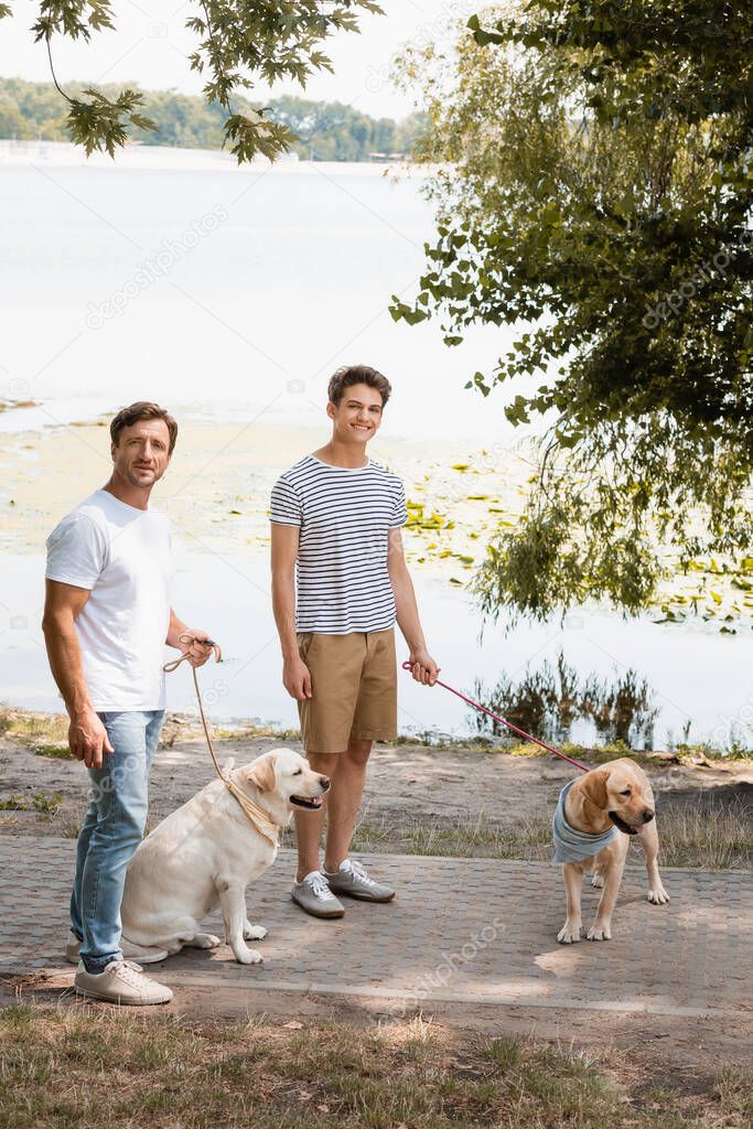father and son holding leashes while standing with golden retrievers near lake 