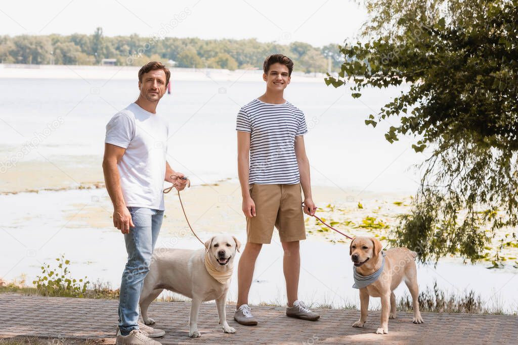 father and teenager son holding leashes and standing with golden retrievers near lake 