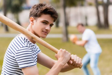 selective focus of teenager son with softball bat looking at camera while playing baseball with father in park clipart