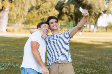 Genç oğlan yeşil parkta selfie çekerken babasına sarılıyor. 