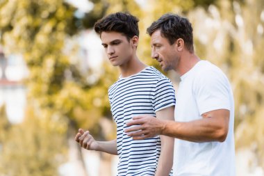 father gesturing and talking with teenager son in park  clipart