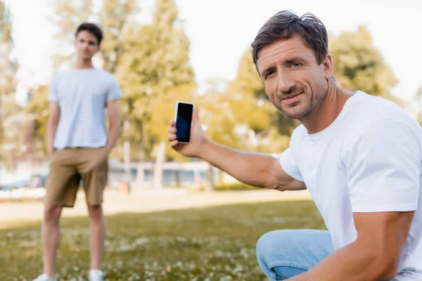 在公园里 父亲拿着带有空白屏幕的智能手机 接近十几岁的儿子时的选择焦点 — 图库照片