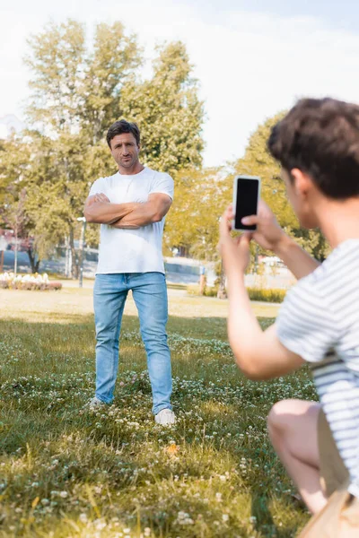 当男孩在公园拍照和拿着智能手机时 男人交叉双臂站在一边的选择性焦点 — 图库照片