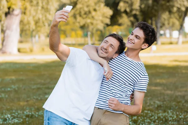 teenager son hugging father while taking selfie in park