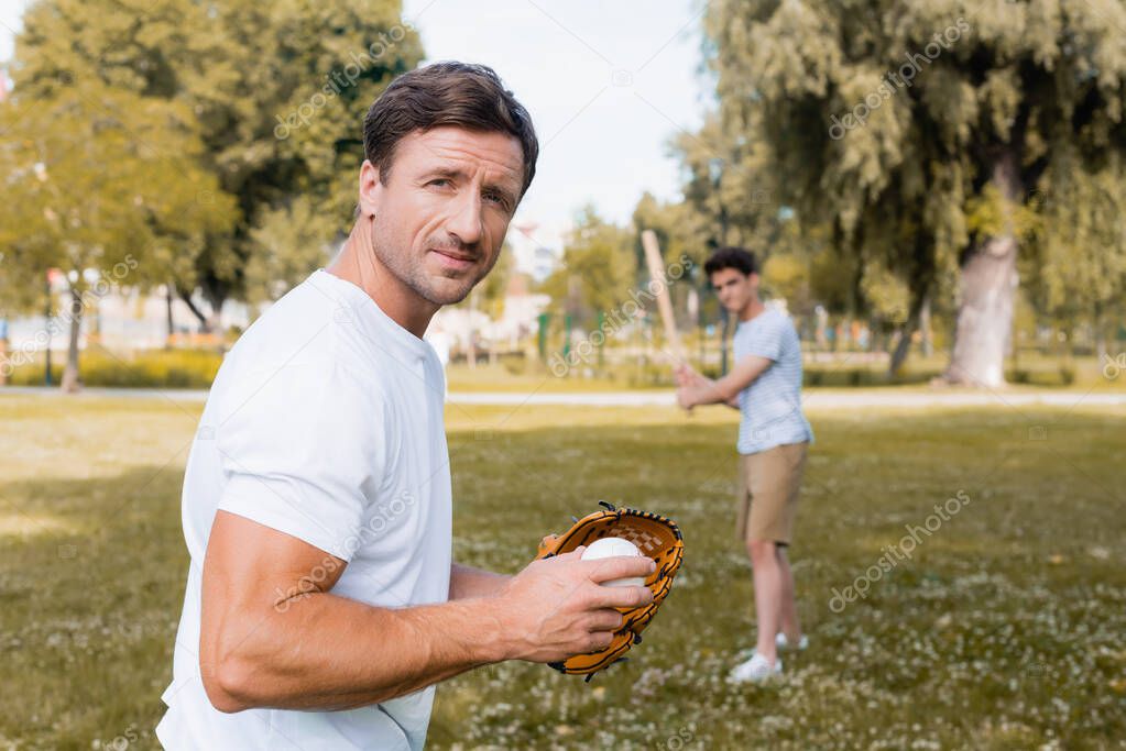 selective focus of man in leather glove looking at camera and holding ball playing baseball with teenager son in park 