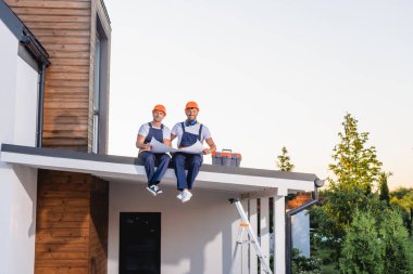 Binanın çatısındaki alet çantasının yanında plan tutarken kameraya bakan inşaatçılar 