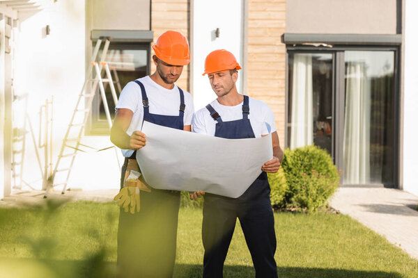 Selective focus of builders in overalls and hardhats looking at blueprint near building 