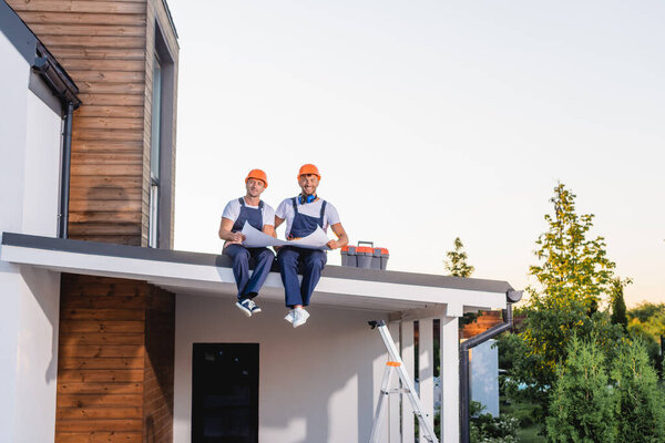 Builders looking at camera while holding blueprint near toolbox on roof of building 