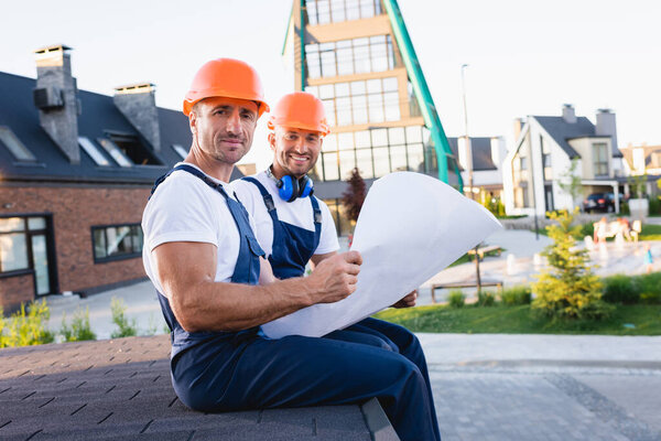 Workmen in overalls holding blueprint on roof of building on urban street 