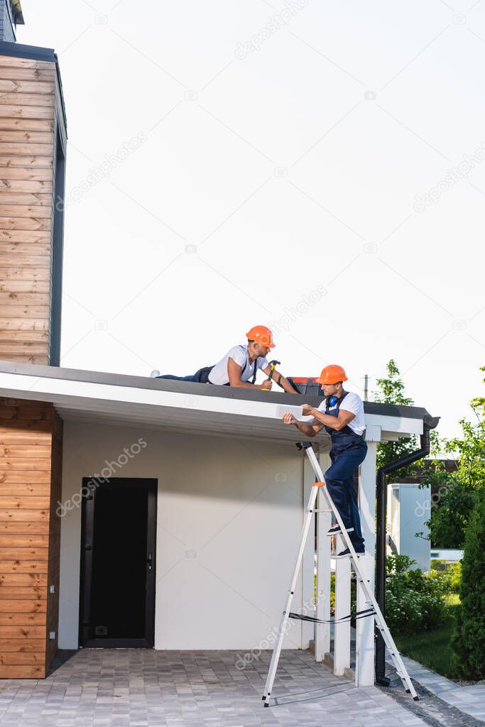 Builder on ladder holding digital tablet near colleague with hammer on roof of house 