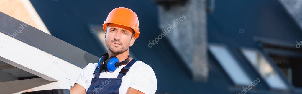 Panoramic shot of builder in hardhat looking at camera near roof of building 