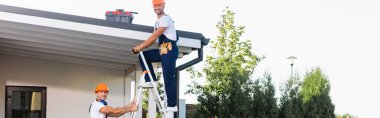 Horizontal crop of builders looking at camera while using ladder near building   clipart