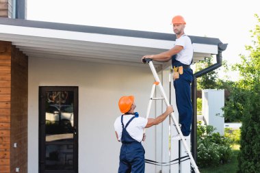 Handymen in hardhats and uniform using ladder near roof of building  clipart