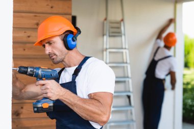 Kask ve üniformalı tamircinin seçici odak noktası bina ön cephesinde elektrikli tornavida kullanıyor. 