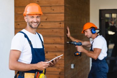 Meslektaşı binanın ön cephesinde elektrikli tornavida kullanırken, inşaatçının dijital tableti tutmasının seçici odak noktası 