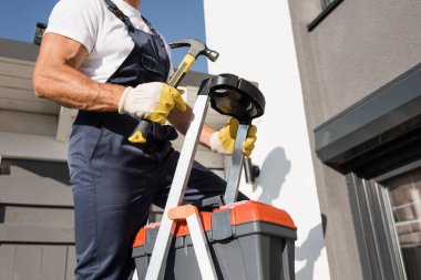 Cropped view of workman with hammer standing on ladder with toolbox near building  clipart