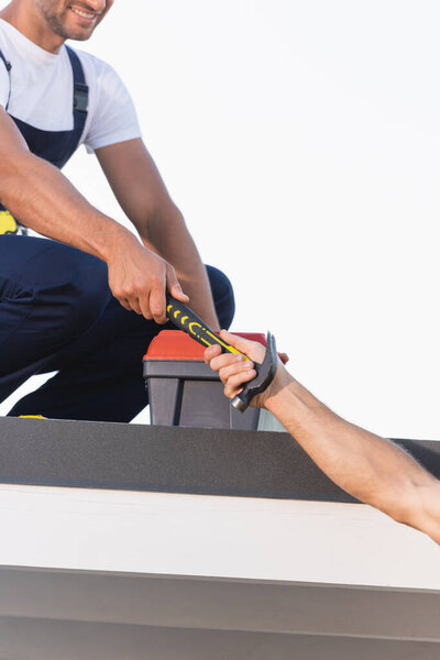 Cropped view of builder giving hammer to colleague near toolbox on roof of building isolated on white 