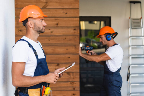 Selective focus of builder in tool belt and workwear holding digital tablet near colleague and house 