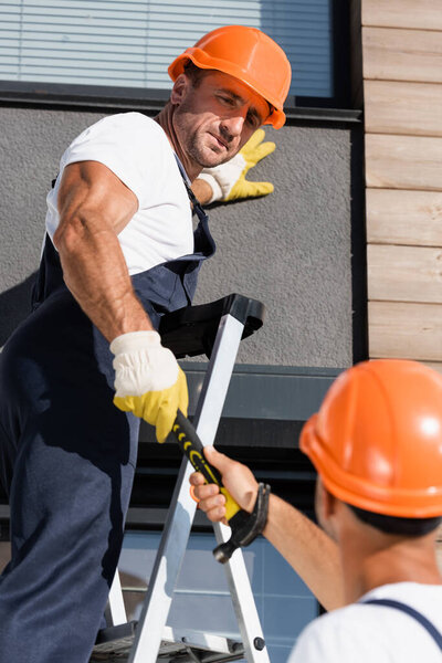 Selective focus of builder giving hammer to colleague on ladder near facade of building 