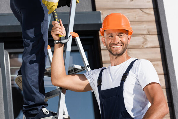 Selective focus of builder looking at camera while giving hammer to colleague on ladder near house 