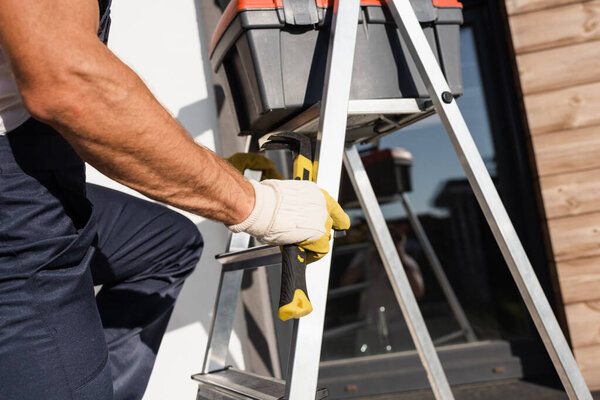 Cropped view of builder in gloves holding hammer near toolbox on ladder outdoors 