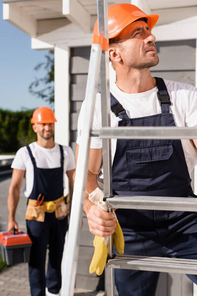 Selective focus of builder in hardhat standing on ladder near colleague and building 