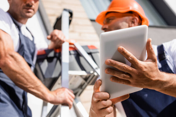 Selective focus of handymen using digital tablet while working with ladder near building 
