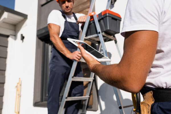 Selective focus of manual worker holding digital tablet beside colleague on ladder and building outdoors 