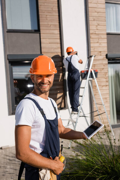 Selective focus of builder holding digital tablet while colleague working on ladder near building 