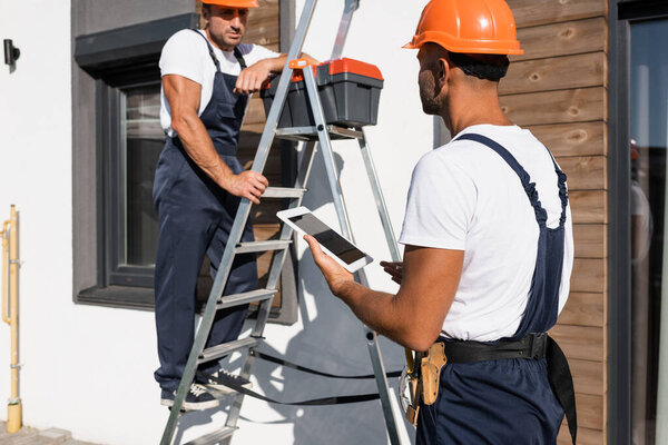 Selective focus of handyman with digital tablet standing beside colleague in uniform on ladder and house 