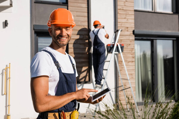 Selective focus of builder pointing with finger at digital tablet while colleague working near building at background 