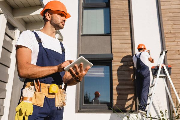 Selective focus of builder in uniform and tool belt using digital tablet while colleague working on ladder beside building  