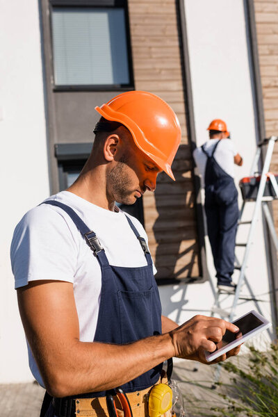 Selective focus of builder in hardhat and tool belt using digital tablet near building outdoors 