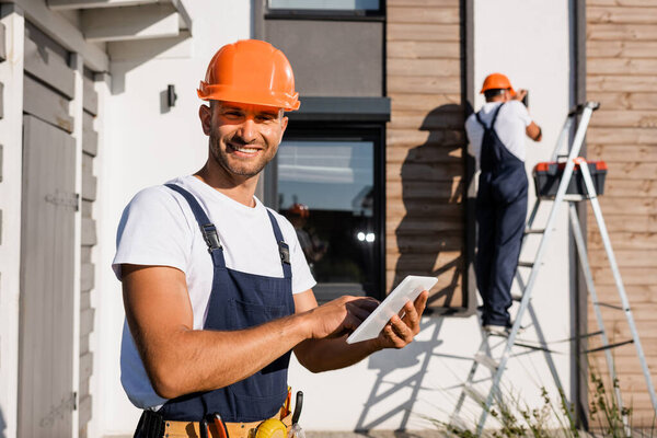 Селективный фокус строителя с цифровым планшетом, смотрящим в камеру, в то время как коллега работает над лестницей возле дома на заднем плане 