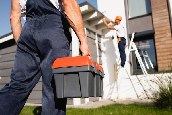 Selective focus of builder holding toolbox while colleague working near house at background 