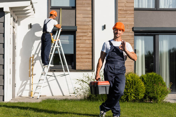 Selective focus of handyman with toolbox showing ok at camera while standing on lawn near building 