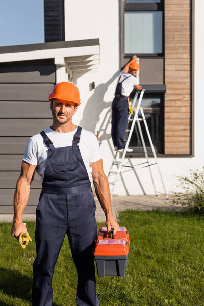 Selective focus of builder with toolbox and gloves looking at camera on lawn near house 